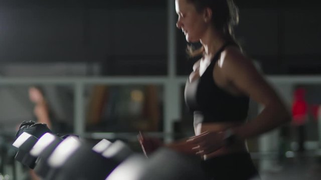 Portrait of a pretty athletic brown-haired woman with a ponytail on her head and a fitness bracelet, engaged in physical culture on a treadmill in a dark room of the gym. Motivation for a healthy