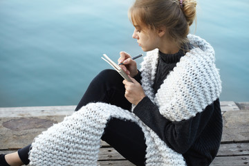 pretty young blonde woman wearing scarf sitting on wooden pier writing notes on notebook with pen