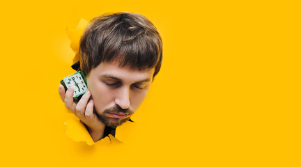 A young guy peers through a torn hole in yellow paper and sleepily holds an green alarm clock near...