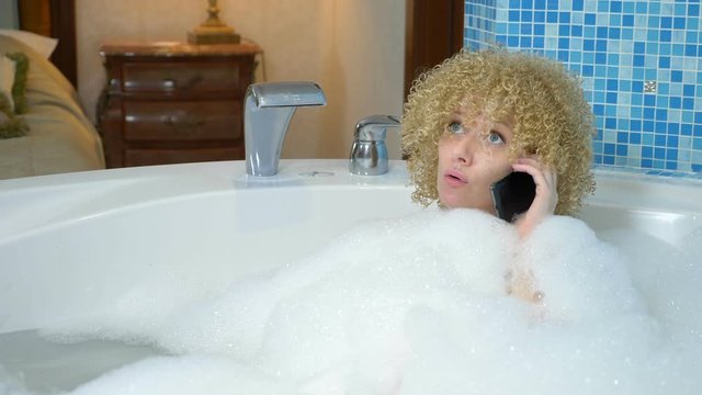 Cute blonde girl in the bathtub with foam dropped the phone into the water. and trying to get the fallen phone out of the water.