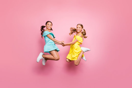 Full body profile photo of two small sisters jumping high together last studying day holding hands best friends wear blue yellow dresses isolated pink color background