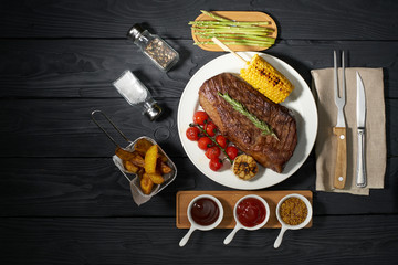 grilled steak on a plate with asparagus and tomatoes, French fries and country potatoes. Three different sauces: ketchup, mustard and barbecue on a dark wooden background, top view, horizontal photo