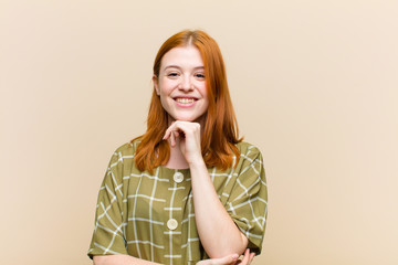 young red head pretty woman looking happy and smiling with hand on chin, wondering or asking a...