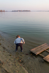 Fototapeta premium Little kid boy on a wooden pier on a lake, a river looks into the distance at the horizon