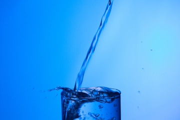 Chorro de agua llenando y rebosando un vaso de agua de cristal o plástico transparente sobre fondo azul
