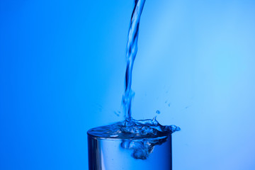 Chorro de agua llenando y rebosando un vaso de agua de cristal o plástico transparente sobre fondo azul
