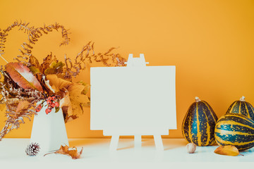 Empty blank canvas on a stand, decorative striped pumpkins, vase with bouquet of falling leaves and fern on orange wall background. Autumn natural home interior decor. Eco, simple style. Copy space.