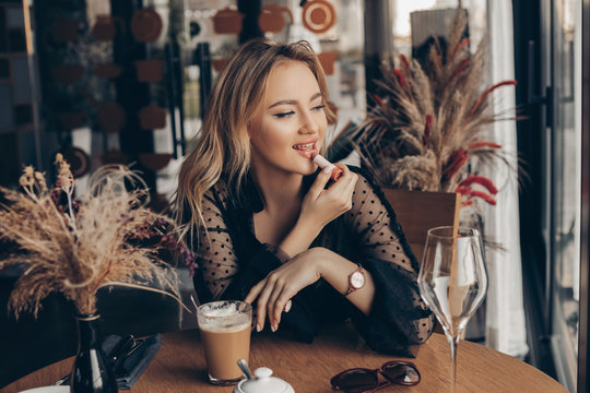 Fashionable beautiful girl applies lip make-up with peach lipstick while sitting at a table in an elegant interior. fashion portrait stylish curly blonde girl in a black jacket. Caucasian young woman 