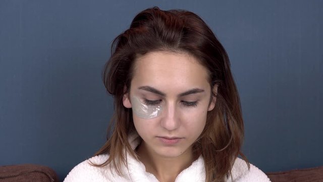 Young woman puts patches on her face. Blue hydrogel patches for rejuvenating the skin of the face. Girl sitting in a white coat. Close up