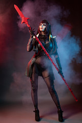 Beautiful leggy busty cosplay girl wearing an erotic leather costume posing with a fake spear on a red background in theatrical smoke.