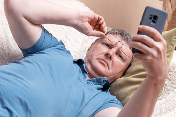 A man with glasses looks at a smartphone while lying in a cozy sofa. Middle-aged businessman checks...