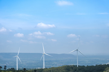 The big white wind turbine for spinning electricity sees large mountains far away, with clouds and blue skies above the mountains.