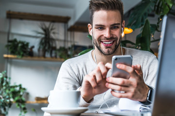 Portrait of smiling man with earphones sitting at coffee shop and using mobile phone