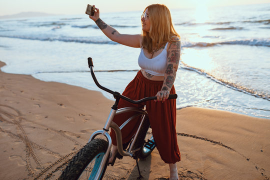 Cheerful chubby woman taking selfie on smartphone while resting with bicycle at sunny shore
