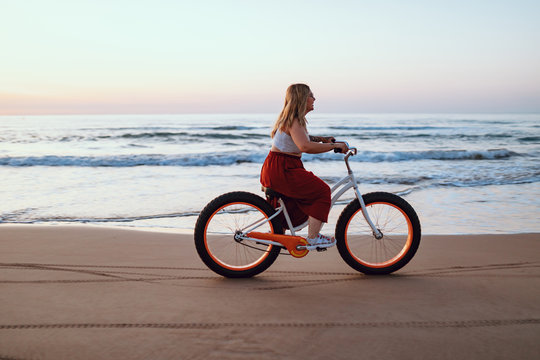 Side view of chubby modern happy woman in bright skirt riding bicycle along scenic ocean shore in twilight
