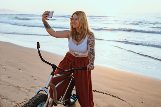 Smiling content tattooed woman in glasses taking picture on smartphone while standing with bike at peaceful beach