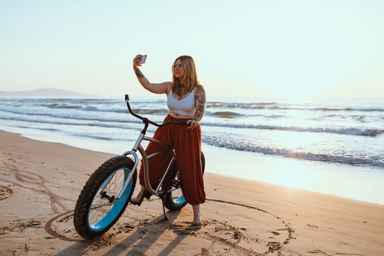 Cheerful chubby woman taking selfie on smartphone while resting with bicycle at sunny shore