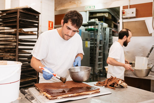 Concentrated pastry cook smearing chocolate topping on tasty cake while standing near table in bakery