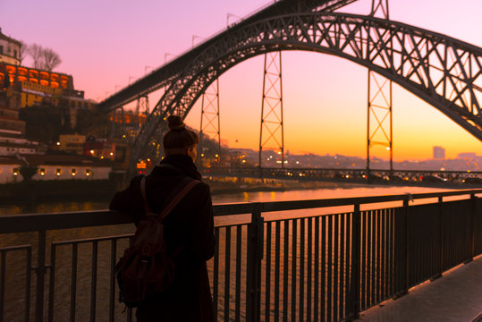 Back view of female tourist standing near city embankment near bridge looking away during sunset in Porto, Portugal