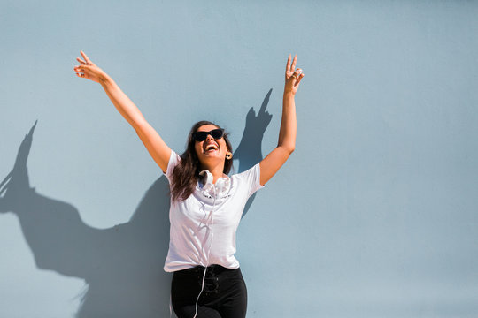 Carefree beautiful woman in casual outfit and headphones on neck standing with hands up beside blue wall of building on city street