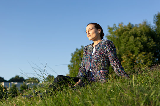 businesswoman in elegant jacket touching neck and looking away while sitting on grass against bushes in park and relaxing