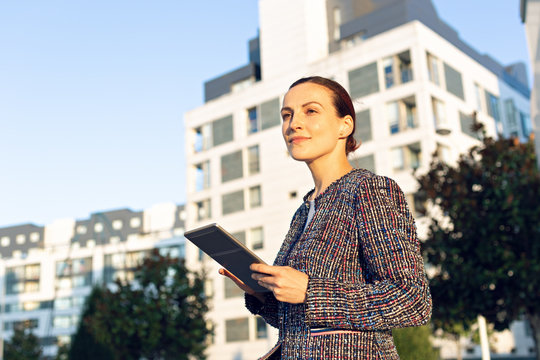 Serious businesswoman in elegant jacket browsing tablet and looking away while standing on city street on sunny day