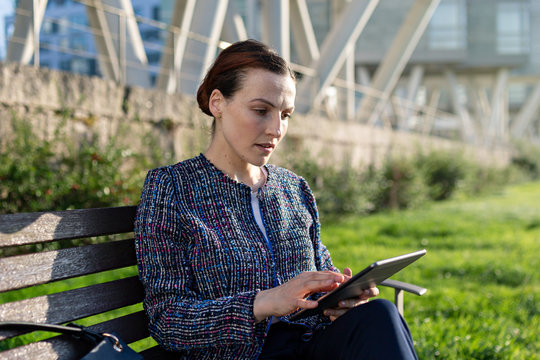Serious female entrepreneur in elegant jacket frowning and using tablet while sitting on bench on sunny day in city park