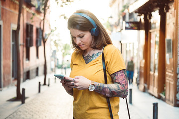Attentive hipster tattooed woman in headphones using smartphone while standing on sunny city street