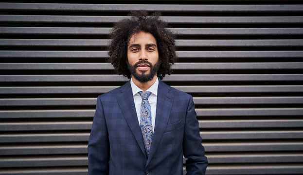 Confident ethnic employee with afro hair and beard standing near striped wall and looking at camera