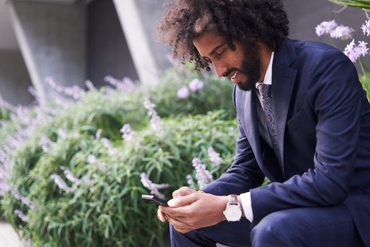 Joyful ethnic man in suit sitting outside and messaging