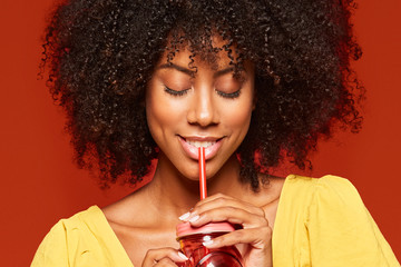 Close-up view of dreamy young African American lady with curly hair holding red jar with straw and enjoying beverage on red background