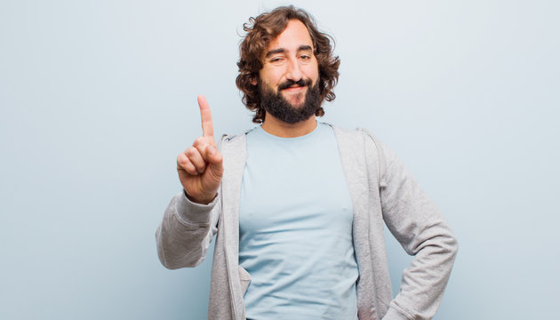 young bearded crazy man smiling and looking friendly, showing number one or first with hand forward, counting down against flat color wall