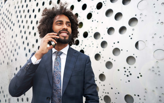 Confident African American businessman in formal suit standing near wall with many round holes and talking on smartphone while looking away