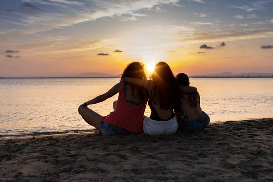 Back view of bronzed female friends hugging and relaxing on beach at sunset in summer in back lit