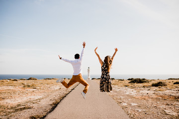 Back view of content lovers bouncing on pathway to lighthouse and sea in summertime