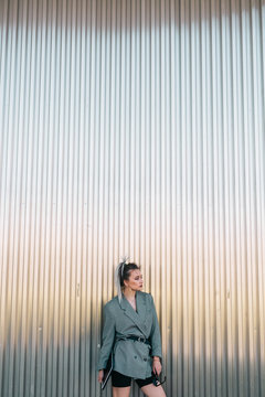 Trendy businesswoman holding tablet and sunglasses leaning metal wall and looking away with questioning expression