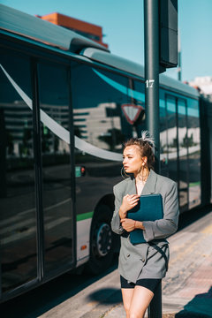 Trendy businesswoman holding tablet wearing suit and sunglasses leaning on bus stop and looking away