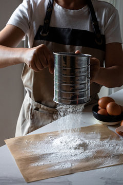 Crop person in apron standing by table sifting flour on baking paper with stainless sifter for making dough