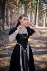 Portrait of a young, beautiful girl in a black medieval dress standing in a pine forest.