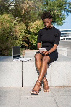 Concentrated African American woman in elegant black dress using mobile phone near a laptop while relaxing on pavement on street