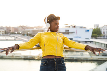 Content African American woman in trendy jacket listening to music in headphones while leaning on glass balcony and looking away on blurred background