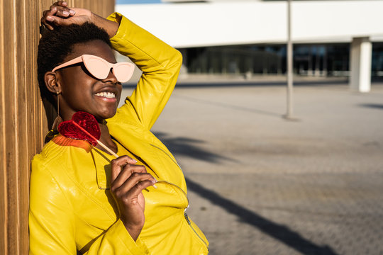 Side view of trendy African American woman in sunglasses in yellow jacket enjoying heart shaped lollipop leaning by wooden fence