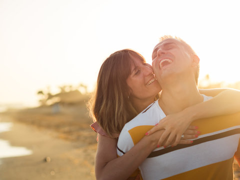 Full length cheerful adult male giving piggyback ride to smiling female while standing on wet sand near waving sea on resort and looking at camera