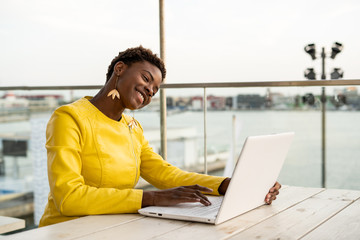 Black African American woman in yellow jacket using laptop web cam at wooden desk in city on blurred background