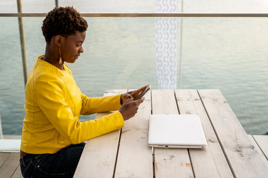 Concentrated African American female messaging smartphone while relaxing at wooden table