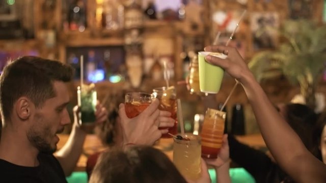 Handheld shot of group of cheerful young people of different ethnicities holding glasses with cocktails while dancing at party at bar or nightclub