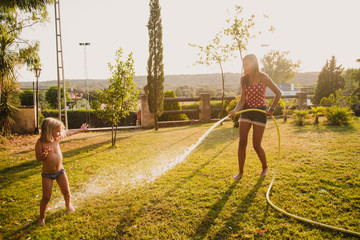 Full body cheerful teenager hosing little girl with clean water while having fun on lawn in yard together