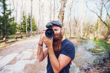 Modern bearded redhead man in cap taking photo with camera while standing on road surrounded with leafless trees in colorful woods on sunny autumn day