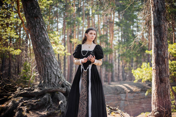 Fototapeta premium Young, beautiful girl in a black medieval dress with a steel rose in hand, walking through the forest.