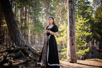 Fototapeta premium Young, beautiful girl in a black medieval dress with a steel rose in hand, walking through the forest.
